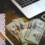 A workspace setup with dollar bills, notepad, and laptop on a wooden desk.