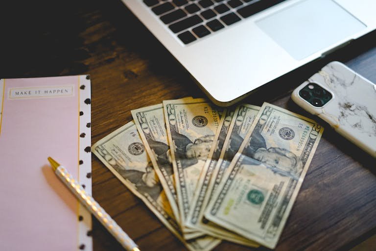 A workspace setup with dollar bills, notepad, and laptop on a wooden desk.
