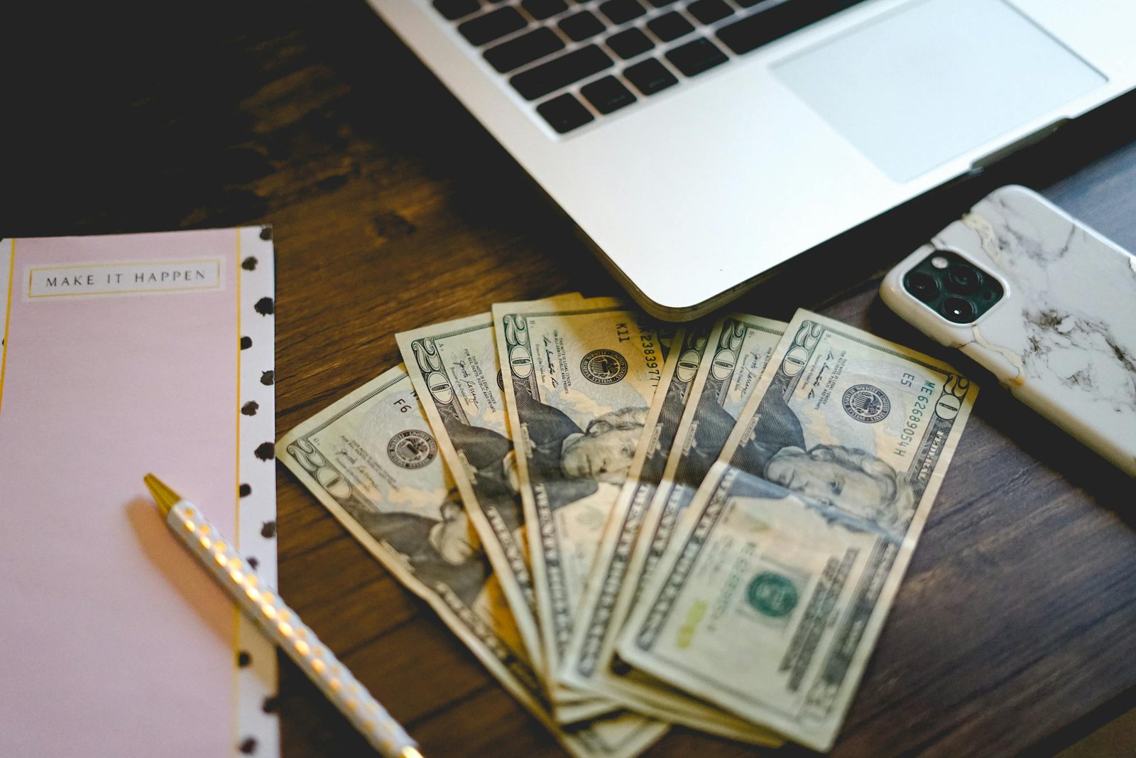 A workspace setup with dollar bills, notepad, and laptop on a wooden desk.